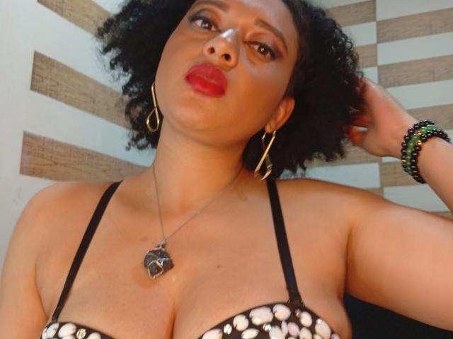 VioletaSwitch with curly black hair, red lips, gold hoop earrings, black strap dress with white beads, heart necklace, and green bracelet, looking up with a pouty expression.