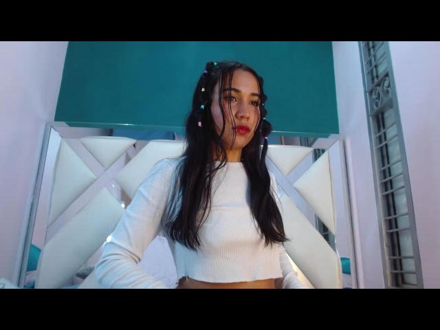 KateWendys stands in a room with a white, diamond-patterned headboard. She has long, dark hair in pigtails with small, colorful hair ties. She wears a white, long-sleeve crop top, exposing her midriff. Her face is slightly turned to the side, with red lipstick and a neutral expression. The green wall in the background contrasts with the white headboard. The photo has a slight blue tint, emphasizing the green wall. KateWendys is centered in the frame, with her head slightly above the headboard.