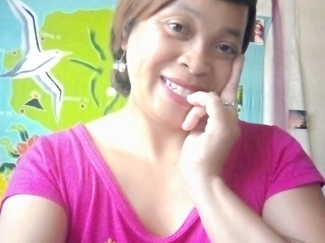 Rahel69 with short brown hair, smiling, wearing a pink shirt, touching her chin with her left hand, showing a ring on her finger, and wearing small earrings.