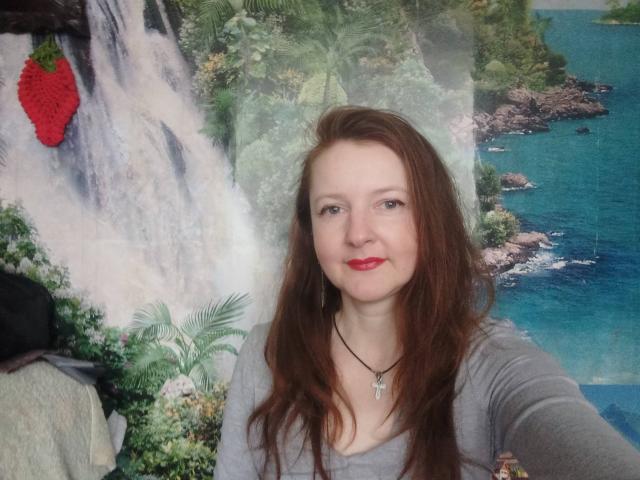 MeganPirs has red hair, red lipstick, gray top, black necklace with cross, fair skin, and is smiling at the camera, with a red strawberry sticker on the left.