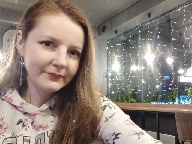 MeganPirs with long red hair, light skin, red lipstick, floral shirt, eyeliner, and earrings, smiling softly, sitting in a brightly lit room with string lights in the background.