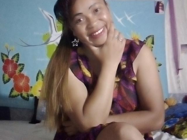 Rahel69 has tan skin, long brown hair, and is smiling with her hand on her cheek, wearing a purple floral dress and star earrings, showing her tits slightly.