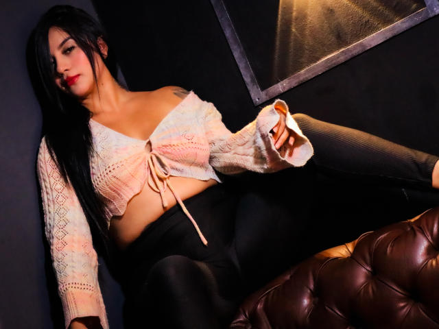 LiaHoffman with long black hair, wearing a pink off-shoulder crop top and black leggings, sitting on a brown tufted leather couch, holding a cigarette. Boobs, ass.