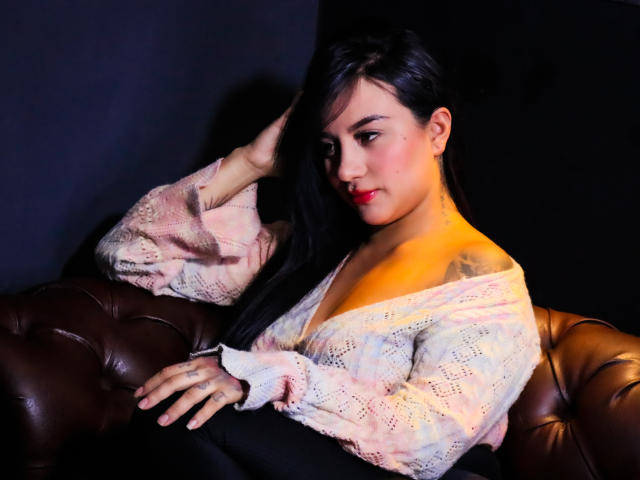 LiaHoffman with long black hair, pink lace-off-shoulder top, and red lipstick, sitting on brown leather couch, hand in hair, black background, tattoo visible on right shoulder.