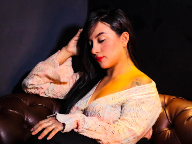 LiaHoffman with long black hair, wearing a pink lace off-shoulder top, resting her hand on a brown leather tufted chair, her red lips and tattooed neck visible.