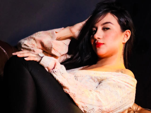 LiaHoffman with long black hair, red lipstick, and a cute smile, wearing a pink off-shoulder sweater and black pants, reclining with one arm behind her head.