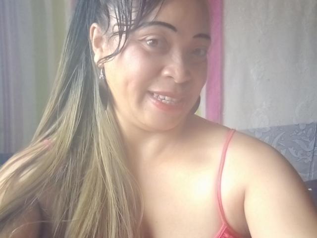 Close-up of Rahel69 with light brown skin, smiling slightly. She has long, straight, dark brown hair with blonde highlights, falling over her right shoulder. She wears a red spaghetti-strap top, exposing her shoulders. She has small, silver earrings and subtle makeup. Her nose is slightly upturned, and she has small, white teeth. The background is blurred with pink and yellow vertical stripes. The light is soft, creating a natural, relaxed look.