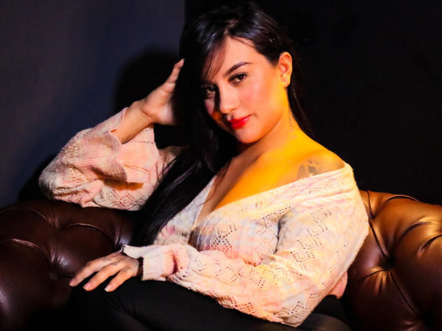 LiaHoffman with long black hair, pink knit top, and brown leather couch, hand in hair, slight smile, red lipstick, visible tattoo on left shoulder.