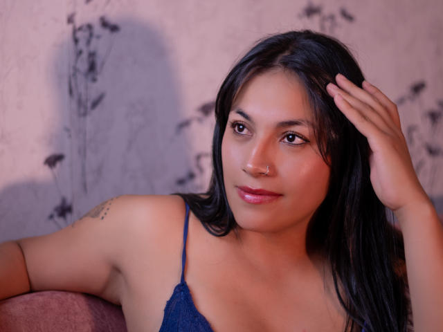 KaitLopez tem a pele marrom clara, cabelo preto e olhos castanhos, vestindo um sutiã de renda azul, com um piercing no nariz e uma tatuagem no ombro esquerdo, e está tocando o cabelo..