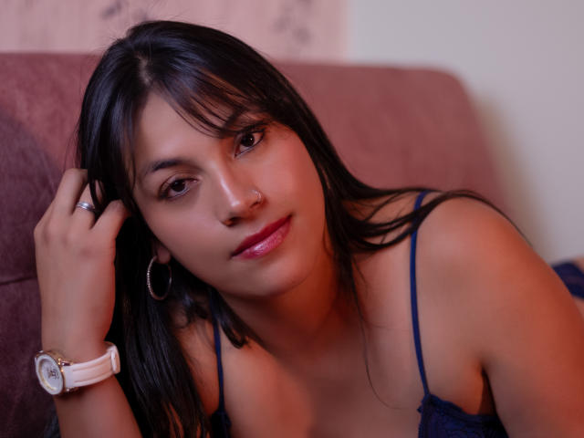 KaitLopez has dark hair, brown eyes, pink lipstick, and a slight smile. She's wearing a blue spaghetti-strap top, a white watch, and silver hoop earrings, touching her hair.