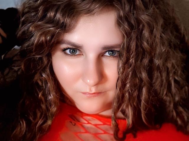 MinistraX has curly brown hair, green eyes, and a slight smile, wearing a red top with a net-like design, showing a subtle nose piercing. 