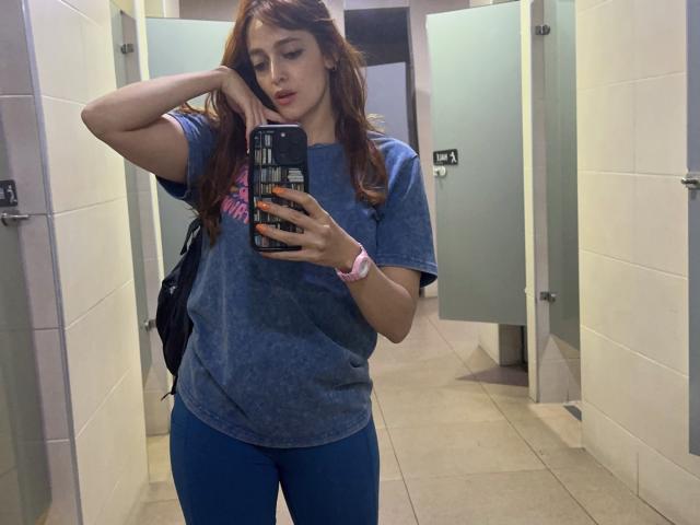 JaneRoa met golvend rood haar, een blauw T-shirt en blauwe broek, die een telefoon vasthoudt, in de badkamer, een spiegelzelfportret, met een roze horloge, een zwarte rugzak, met haar hand in haar haar, en die naar de telefoon kijkt..