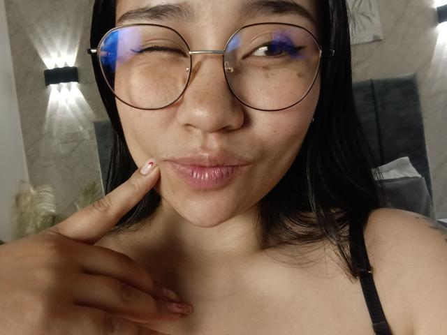 Karollteyes com óculos, piscando, lábios curvados, cabelo preto, pele bronzeada, usando uma alça preta, dedo na bochecha, óculos refletivos, sardas sutis e uma expressão fofa e brincalhona..