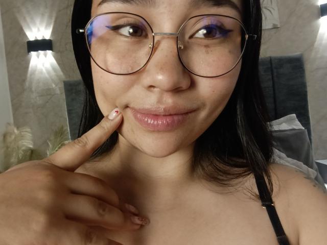 Karollteyes with glasses, purple eyeliner, and a cute smile, touching her chin with her finger, showing her small tits, and pink lips, in a selfie.
