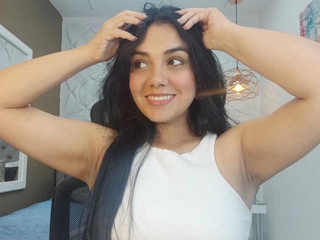 DanMartines with long black hair, smiling, arms raised, hands in hair, wearing a white sleeveless top, showing armpits, brown skin, and looking to the side.