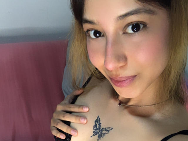 LucyLovelyAngel with light brown hair, black bra, hand on tits, butterfly tattoo, smiling, brown eyes, pink lips, necklace, soft lighting, slightly flushed face, close-up selfie.