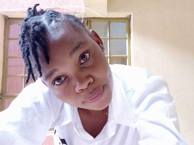 Close-up of PinkyStar with dark brown skin and small, round brown eyes. PinkyStar has short, twisted black hair and is looking directly at the camera with a slight smile. PinkyStar wears a white, button-down shirt. The image is brightly lit, with soft shadows on PinkyStar's face. The background includes a window with yellow-green frames and a pink wall. The composition centers PinkyStar's face, emphasizing their expression and gaze. The texture of PinkyStar's skin is smooth, and the light highlights their facial features.