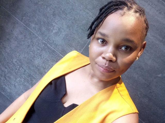 PinkyStar, a Black woman with medium brown skin, is in a close-up selfie. She has small, dark brown eyes and thin, dark brown lips. Her black, twisted hair is pulled back. She wears a bright yellow sleeveless vest over a black top. The vest's yellow color contrasts with the dark background. The image has a slightly angled, top-down perspective. PinkyStar's expression is neutral, with a slight, closed-mouth smile. She has small, gold earrings. The texture of the vest is smooth, while the background is a flat, gray surface.