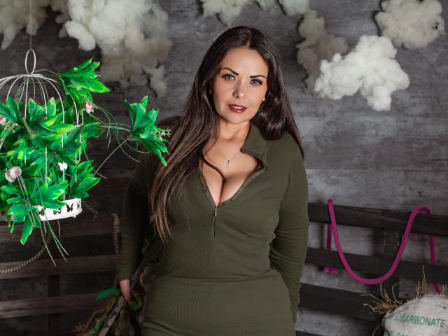 MelissaLeen with long brown hair, green zip-up top showing her big boobs, standing in front of a green plant, gray wall, and pink rope, looking at the camera.