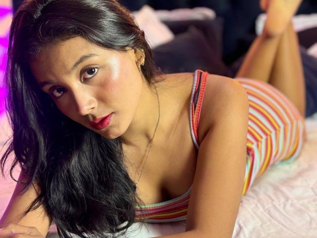 AlinaMonroe, lying on a bed, wearing a striped, tight, sleeveless dress, showing her ass, with her black hair, red lips, and looking at the camera.