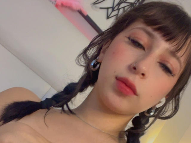 NenaBlack, legata a una sedia blu, indossa un intimo nero con spalline sottili, un morso da pallina, rossetto rosso, con i seni scoperti, le gambe sollevate, un tatuaggio sul fianco, uno sfondo viola e guanti neri..