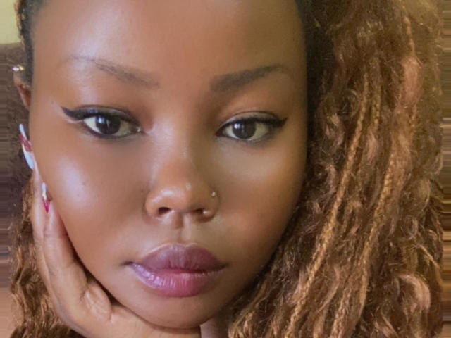 Close-up of AnitaNini with brown, curly hair, resting her chin on her hand. She has medium-dark skin, black almond-shaped eyes with bold black eyeliner, and a small silver nose stud. Her full pink lips are slightly parted. Her long, manicured nails have a white and pink design. The soft, warm lighting highlights her smooth skin. The image has a casual, intimate feel.