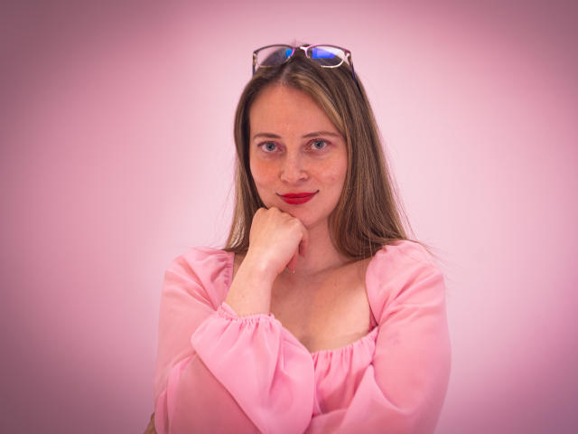 AmbeerWood heeft rode lippenstift, sproeten en draagt een roze blouse, waarbij haar hand onder haar kin rust. Ze heeft blauwe ogen, bruin haar en haar zonnebril zit op haar hoofd..