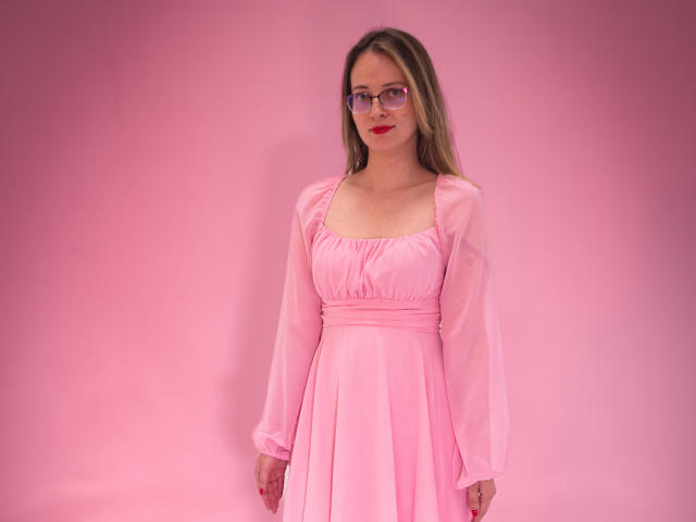 AmbeerWood com óculos, vestido rosa e batom vermelho, em pé, mostrando os seios, fundo rosa, cabelo castanho comprido, fofa, iluminação suave, corpo inteiro, sorriso leve..