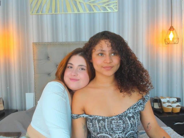 A photo of SofyViky, a curly-haired woman with light brown skin and a small tattoo on her left arm, sitting on a bed. She wears an off-the-shoulder black and white patterned dress. SofyViky has a slight smile and looks to the right. Her arm is around a second woman with pale skin and red hair, wearing a white top. The background includes a beige upholstered headboard, striped wallpaper, and warm orange lights. A decorative yellow leaf pattern is on the wall above. The image has a casual, intimate feel.