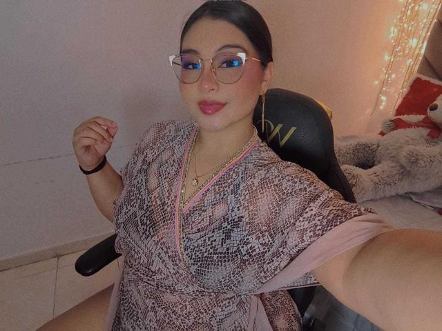 HelueJamil, con ojos azules, usando gafas y un vestido rosa con estampado de serpiente, sentada en una silla, levantando su mano derecha, con lápiz labial rosa, cabello negro recogido en una coleta..