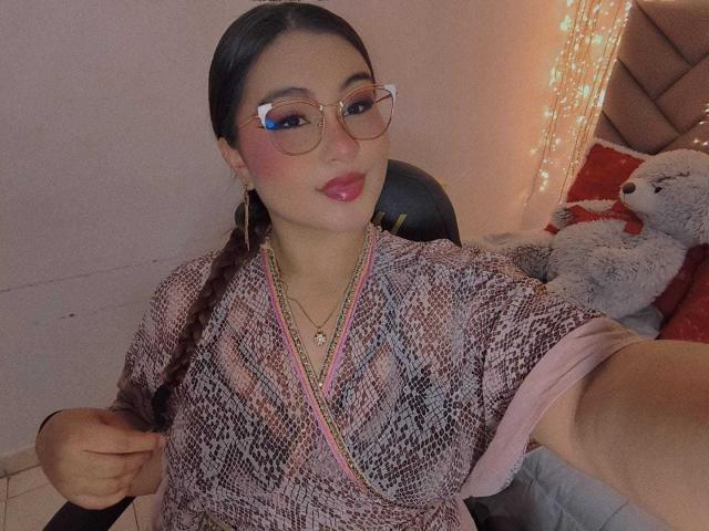 HelueJamil, con lápiz labial rosa, gafas y cabello trenzado, vistiendo una camisa rosa con estampado de serpiente, sosteniendo una manga rosa y un perro de peluche en la cama..