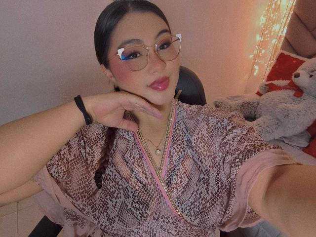 HelueJamil, con piel rosada, cabello negro en trenzas, gafas, lápiz labial rosa, vistiendo una camisa con estampado rosa y marrón, con la mano en la barbilla, un collar, una pulsera negra y un perro de peluche blanco..