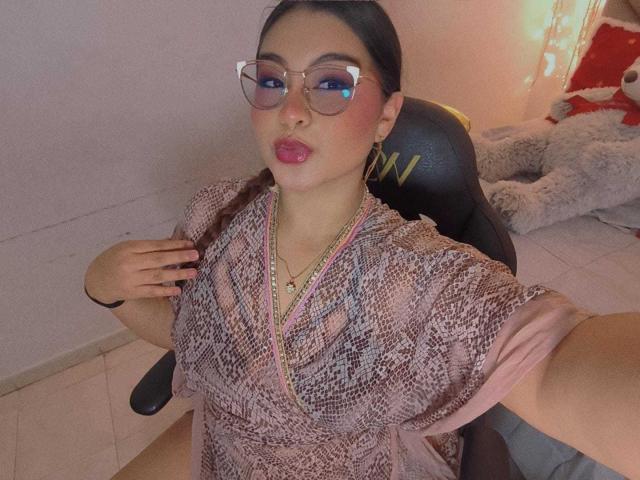 HelueJamil with glasses, pink lipstick, and a snake-pattern dress, showing her tits, sitting on a chair, hand on chest, in a softly lit room.