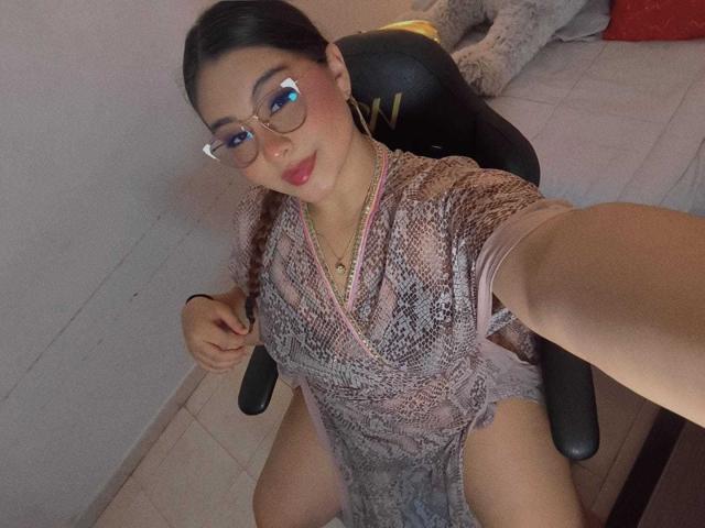 HelueJamil, wearing glasses, pink lace dress, holding braid, sitting on chair, showing bare pussy, smiling, taking selfie, visible tits, cute, light skin, brown hair, red lipstick.