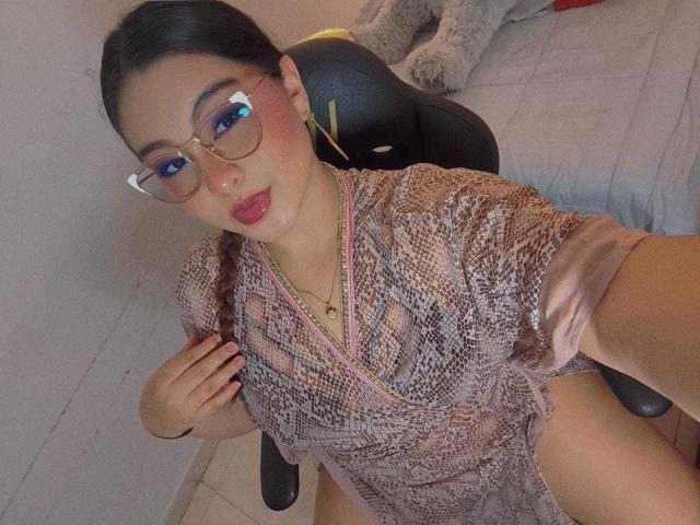 HelueJamil con gafas, vestido de estampado de serpiente rosa, cabello trenzado, lápiz labial rojo, sentada en una silla, mano derecha en el pecho, muslos visibles, cama gris en el fondo, iluminación suave..