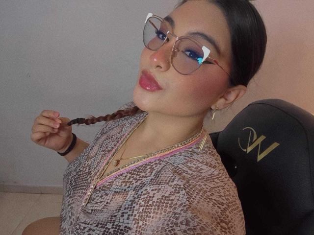 HelueJamil, con gafas, labios rosados y brillantes, sosteniendo una trenza, vistiendo una blusa con estampado de serpiente, sentada en una silla negra con una "W" dorada. Está mirando hacia arriba y sonriendo ligeramente..