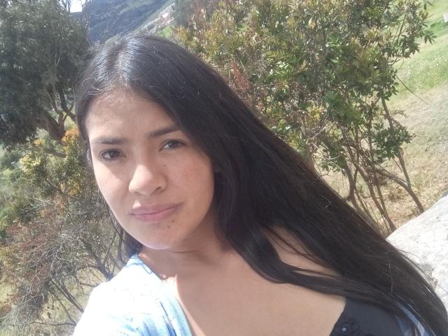 A close-up photo of ArianithaXox with long, straight black hair, light brown skin, and brown eyes. She has a small, subtle smile and wears a blue shirt with a black strap. Her eyebrows are dark and slightly raised. The background shows green trees and a grassy hill. Sunlight illuminates her face and hair. The image is sharp with natural light.
