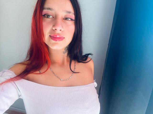 RoxanaSanchezz, con capelli rossi e neri, una maglietta bianca a spalle scoperte, eyeliner rosa e labbra lucide, indossa una collana d'argento e sorride alla fotocamera..