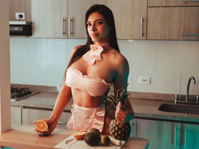 YelineMourine, com seios grandes, vestindo um top e laço rosa de biquíni com babados, segurando um limão, em uma cozinha com uma abacaxi, abacate e limão no balcão..