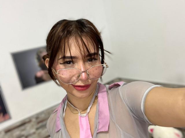 MelannyTaylorr has dark brown hair, pink lipstick, and is wearing sheer glasses with polka dots, a pink collar, and a transparent gray shirt, showing her tits slightly.