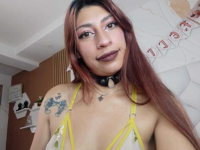 AlisonCold has long red-brown hair, brown eyes, and dark lipstick. She wears a yellow strappy top, black choker, and has tattoos on her left shoulder. Boobs are visible.