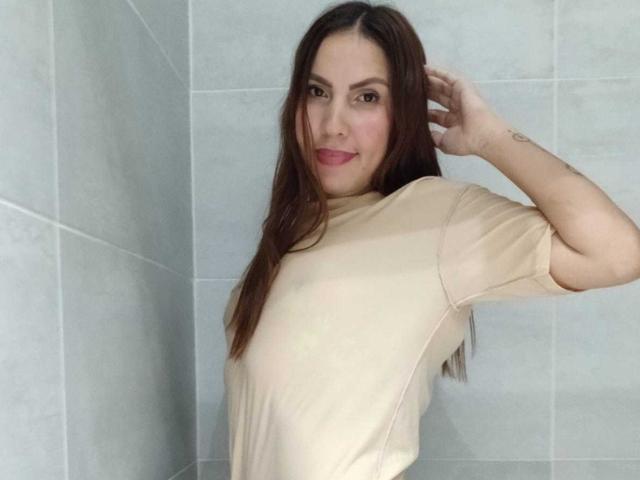 Susan Thomass, met lang bruin haar, lachend, gekleed in een beige shirt, met haar hand in haar haar, toont een kleine tatoeage op haar rechterarm, staand in een betegelde badkamer..