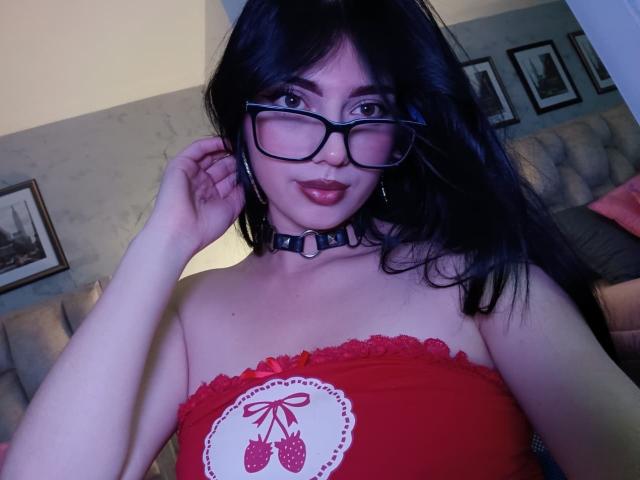 IsabellaaRossi with black hair, glasses, red strapless top with cherry print, black choker, hand adjusting glasses, light skin, full red lips, in a dimly lit room.