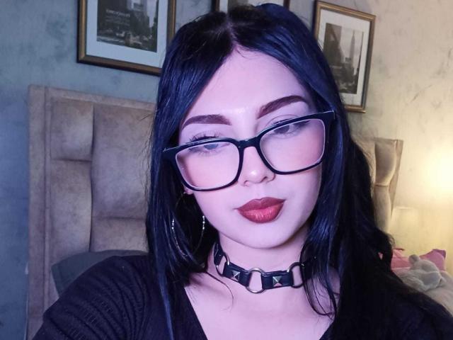Isabella Rossi, com cabelo preto, óculos, batom vermelho, colar preto com anéis de metal e blusa preta, olhando para a câmera com um leve sorriso, sentada na cama..