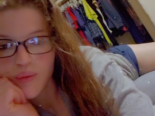 Close-up selfie of Shammie with long, wavy brown hair, wearing black-rimmed glasses and a grey shirt. Shammie's face is partially blurred, resting her head on her hand. She has fair skin and light pink lips. Shammie is lying down with her upper body in the foreground and her arm partially visible. Background includes a closet with colorful clothes. Image has a slight digital filter with small white specks. Shammie's expression is neutral.
