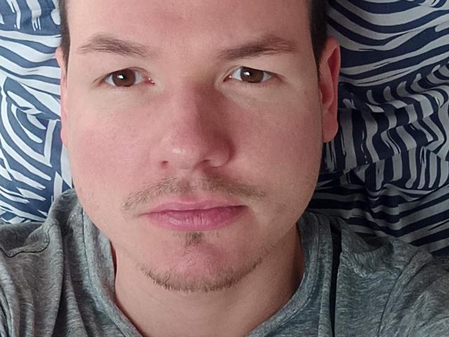 O rosto de ColinFunny, com uma pequena cicatriz acima do olho direito, olhos castanhos, lábios rosados claros, cabelo castanho curto, camisa cinza clara e um bigode/barba fina..