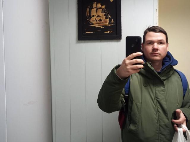Colin, con capelli castano corti, giacca verde, cappuccio blu, che tiene un telefono nero, guarda allo specchio, c'è una parete bianca, un piccolo quadro di una nave, il braccio destro è abbassato, la mano sinistra tiene una borsa bianca..