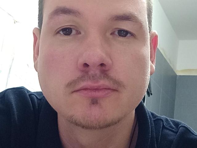 El rostro de ColinFunny, con un bigote corto y barba, con una expresión seria, piel clara, ojos marrones y una camisa negra, en un selfie con un fondo ligeramente borroso..