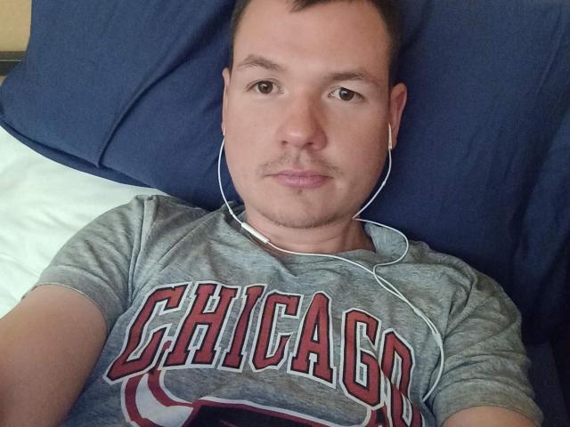 ColinFunny está acostado, vistiendo una camiseta de Chicago, con audífonos blancos puestos, mirando a la cámara con una ligera sonrisa. Tiene un corte de pelo corto y una barba ligera..