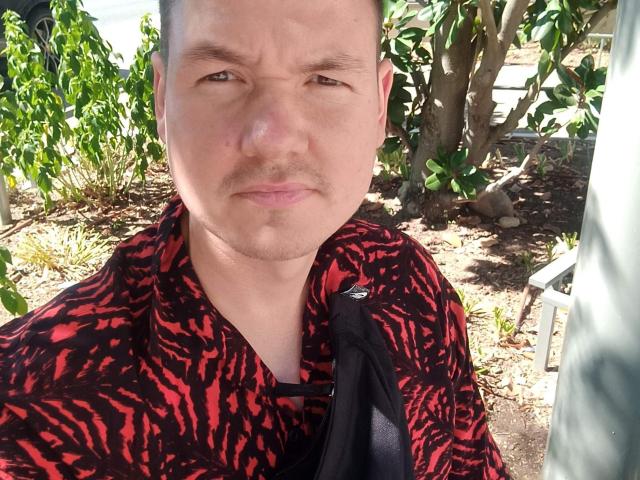 ColinFunny a un visage rond, des cheveux bruns courts, et une chemise à rayures noires et rouges avec un motif zèbre, il a l'air légèrement agacé, avec une petite moustache et un sac noir sur l'épaule..